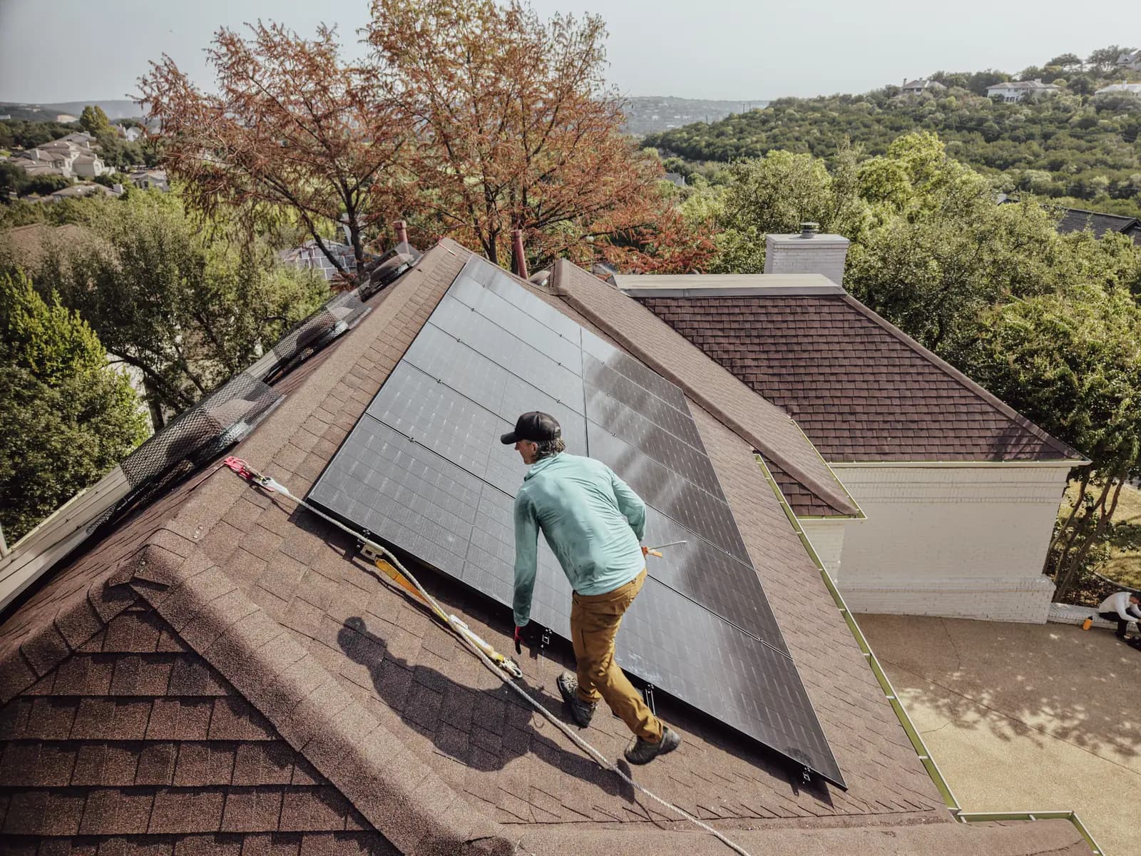 Honest Solar installer walking up a shingle roof beside a newly set solar array, rope line anchored, Austin hill country behind